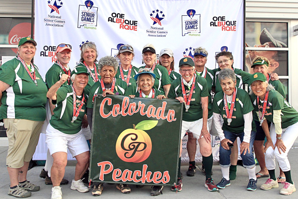 A women's softball team, aged 79-91, smiling together for a team picture outside in their green and white uniforms and red medals, with the center of the front row holding a Colorado Peaches banner, all in front of a white picture banner with assorted logos likely of tournament sponsors.